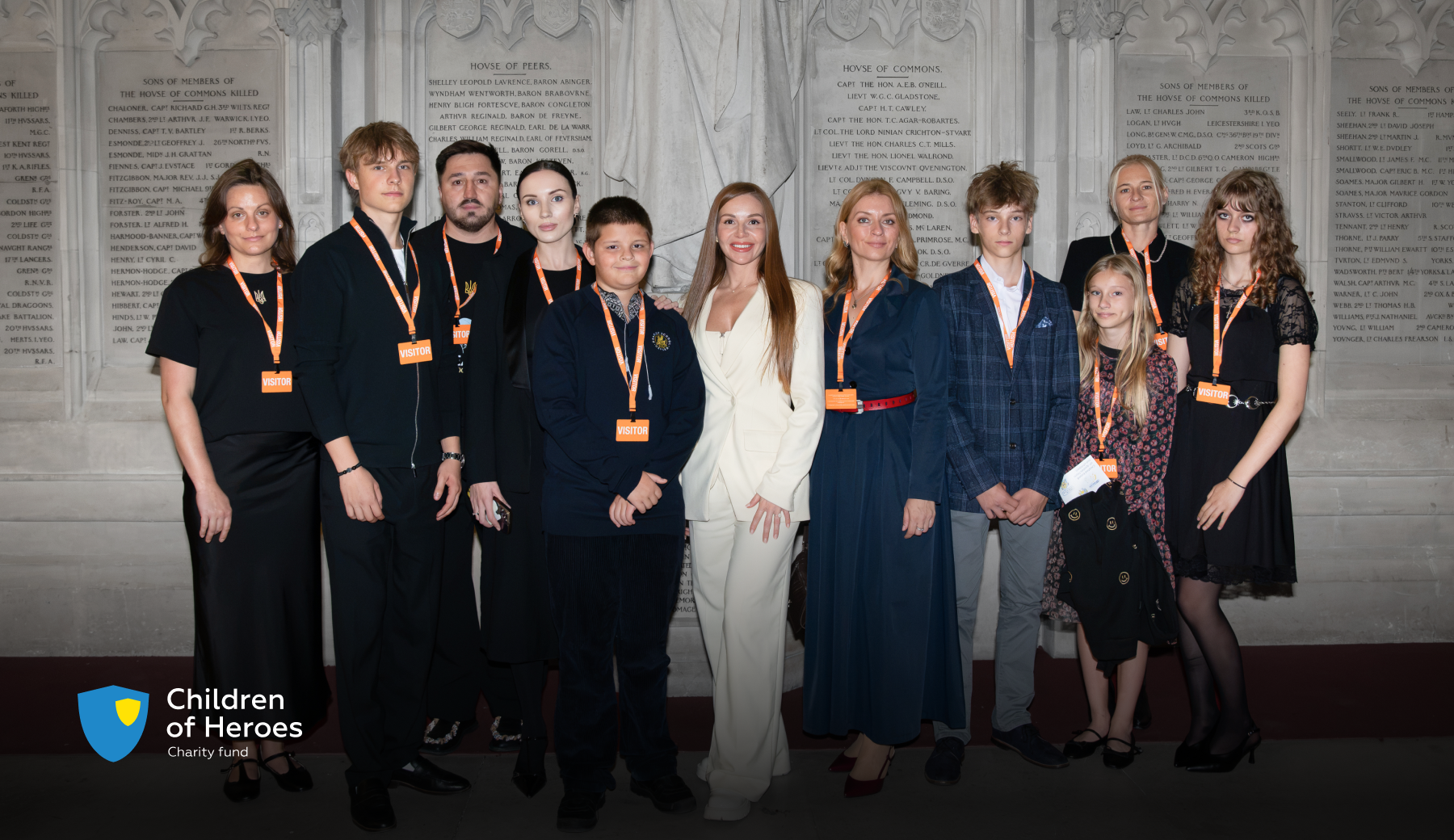 Children of Heroes Fund representatives and Ukrainian families the Fund supports stand together in the House of Lords, wearing orange visitor badges, in front of stone memorial plaques.